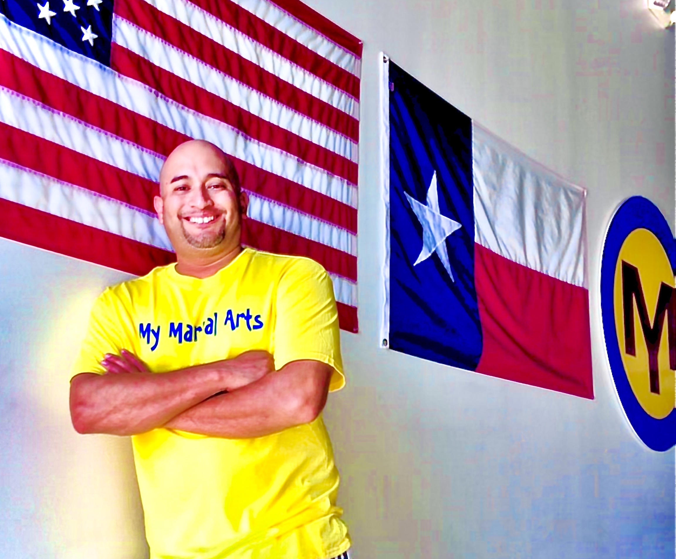 Tony Rios in a yellow My Martial Arts shirt, arms crossed, with the U.S. and Texas flags behind him